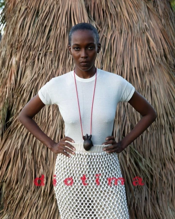 A dark-skinned woman poses in a white shirt and mesh skirt against a backdrop of dried palm fronds in a fashion-style portrait.