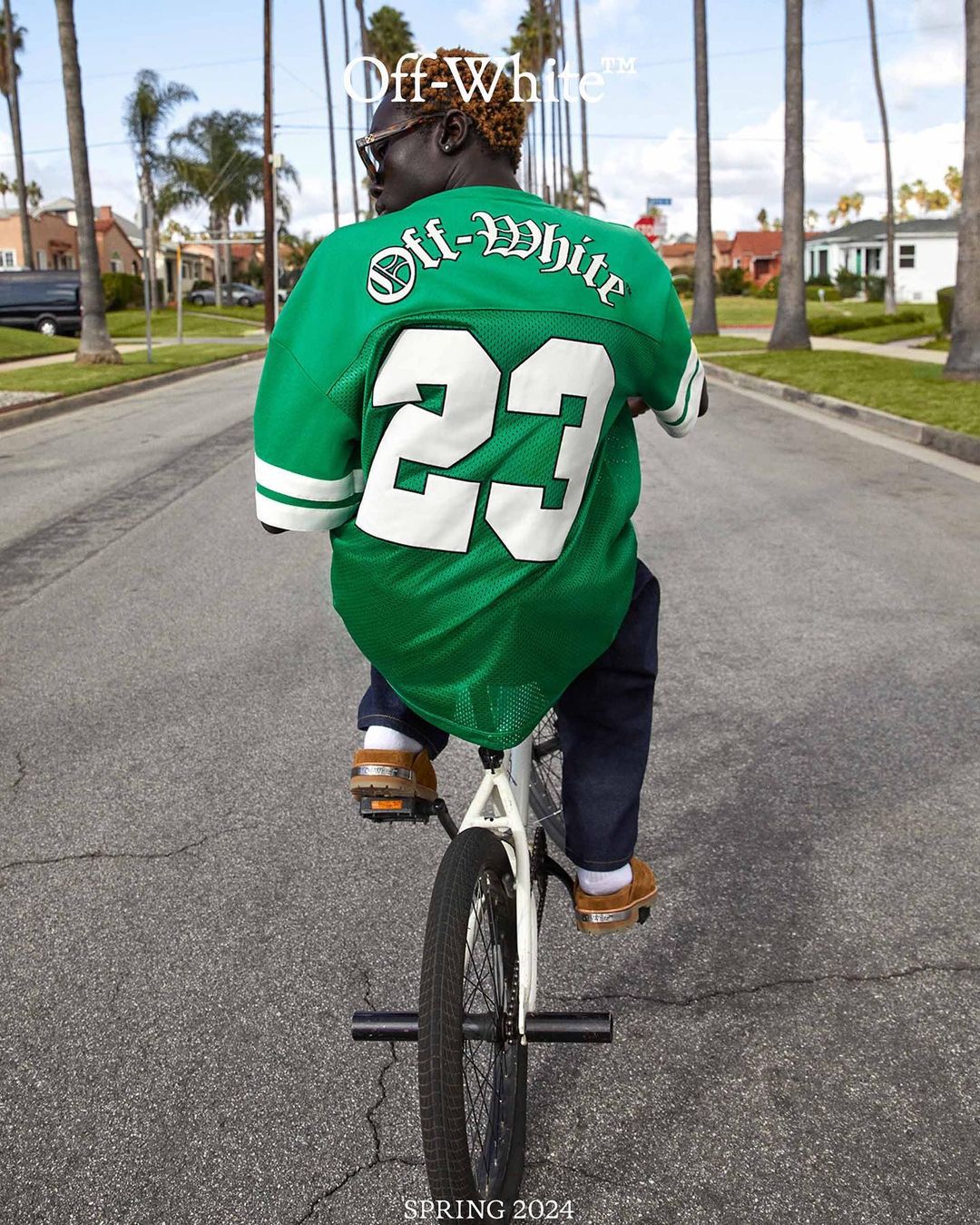An adult rides a BMX bike down a suburban street wearing a green Off-White jersey and jeans, captured in eye-level, medium shot on a sunny day.