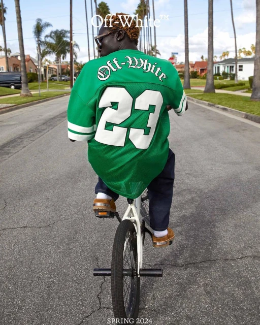 An adult rides a BMX bike down a suburban street wearing a green Off-White jersey and jeans, captured in eye-level, medium shot on a sunny day.