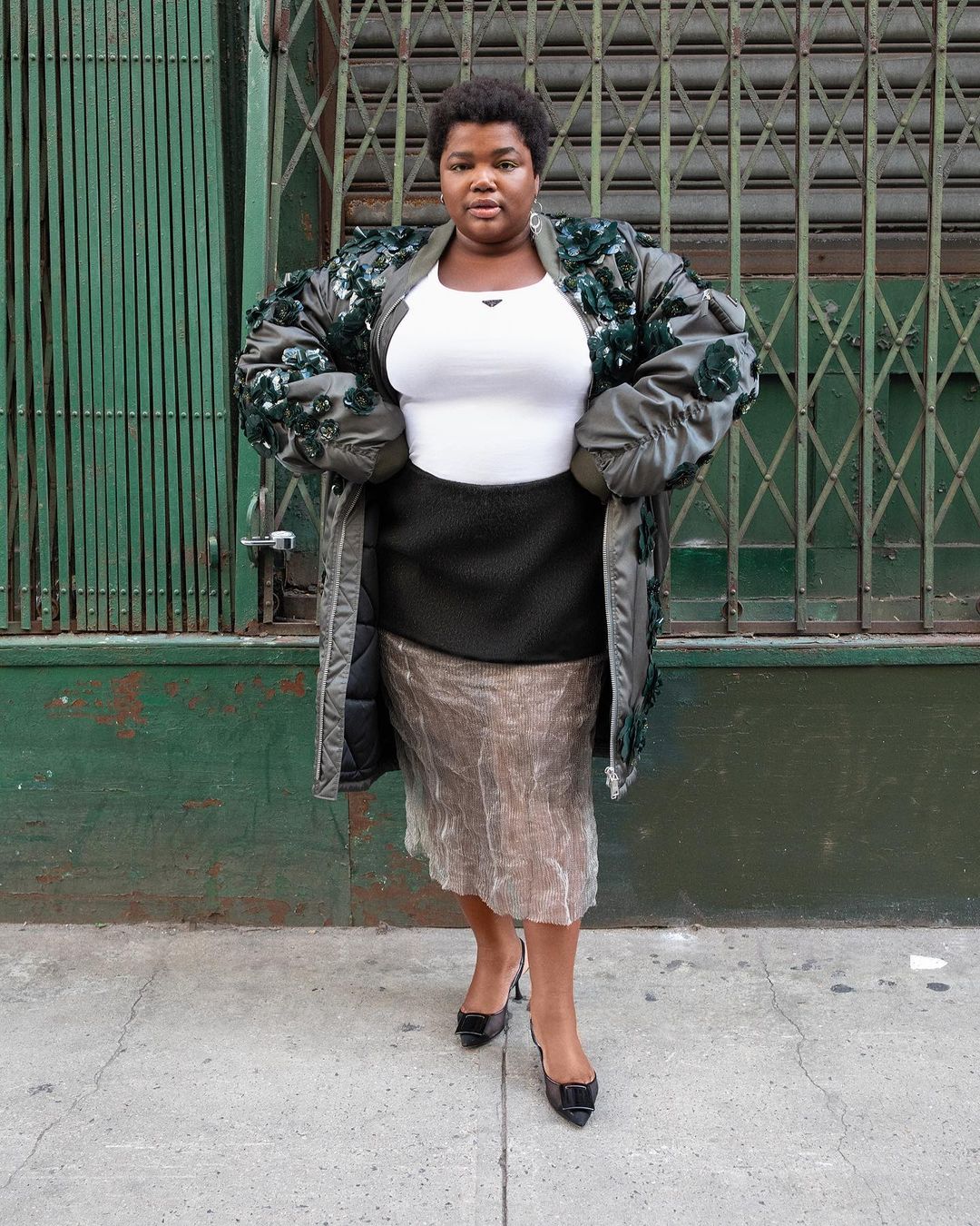 Woman in olive green coat with floral embellishments, black pencil skirt, and patent leather pumps stands against a green metal wall.