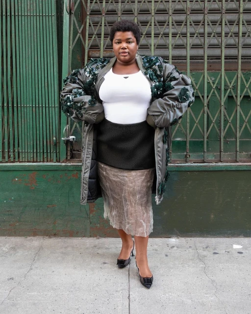 Woman in olive green coat with floral embellishments, black pencil skirt, and patent leather pumps stands against a green metal wall.