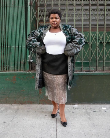 Woman in olive green coat with floral embellishments, black pencil skirt, and patent leather pumps stands against a green metal wall.