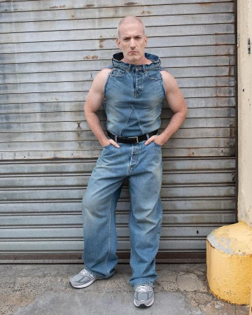 Adult male in a sleeveless denim outfit poses in front of a metal garage door with his hands in his pockets looking at the camera.