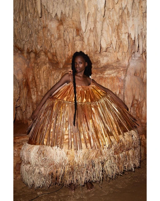 An adult female with long braids poses in a gold and tan dress inside a cave with textured walls and soft lighting, medium shot.