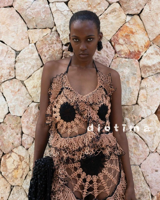 A young Black woman stands in front of a stone wall, wearing a crochet halter dress and carrying a crochet bag in a fashion shot.