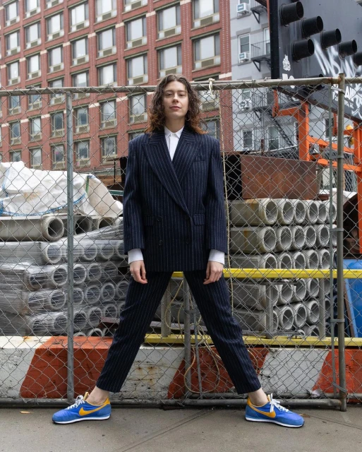 Woman in a striped pantsuit and blue sneakers stands against a construction fence with a building in the background on an overcast day.