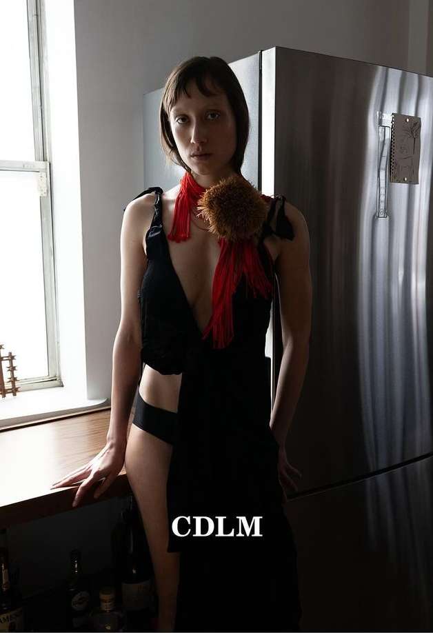 Woman in black dress posing indoors near window and stainless steel refrigerator; she has a red scarf and brown pom-pom accessory.