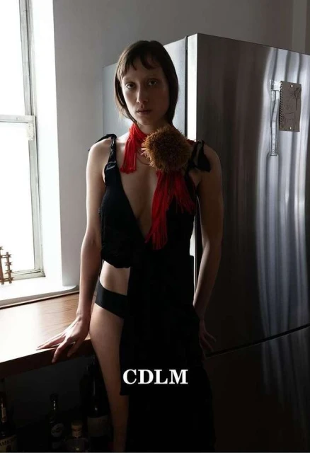 Woman in black dress posing indoors near window and stainless steel refrigerator; she has a red scarf and brown pom-pom accessory.