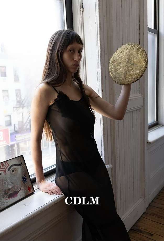 Indoor portrait of a woman holding a gold disc. She's wearing a sheer black dress, standing near a window, and an artwork sits on the ledge.