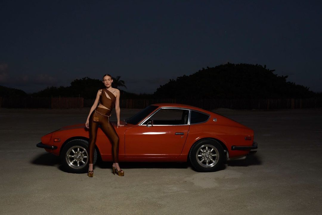 A woman in a brown outfit poses next to an orange vintage car on a beach under the night sky with sparse vegetation in the background.