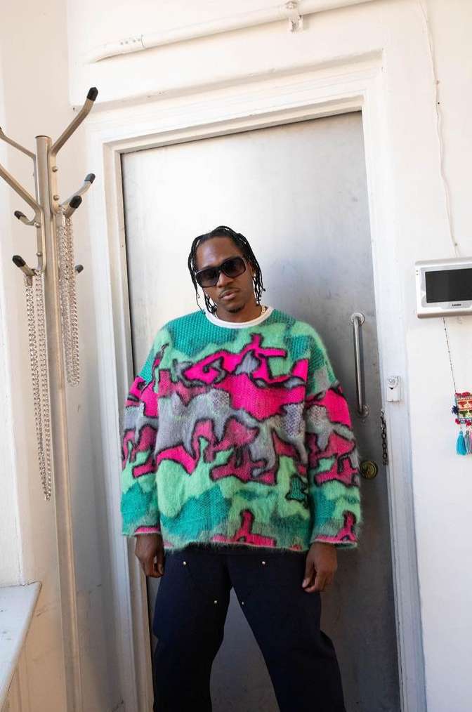 Adult male poses indoors wearing a colorful patterned sweater, dark pants, and sunglasses next to a door with a metal coat rack.