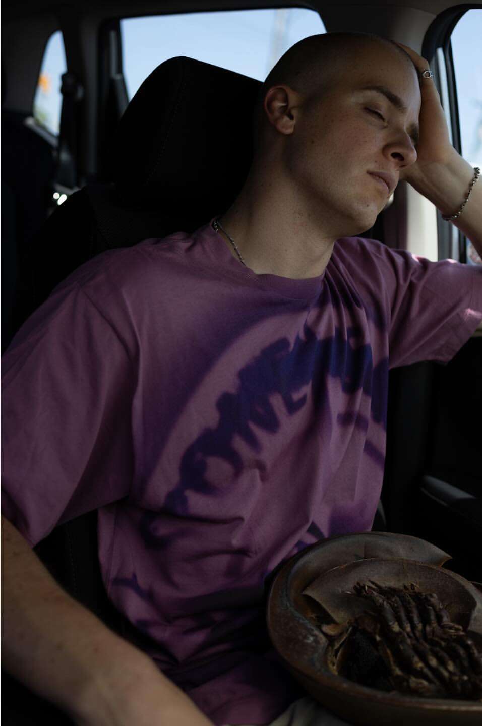 A young adult man is seated inside a car, eyes closed, with his hand on his head, next to a round bowl containing objects.