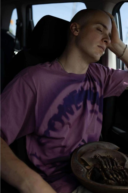 A young adult man is seated inside a car, eyes closed, with his hand on his head, next to a round bowl containing objects.