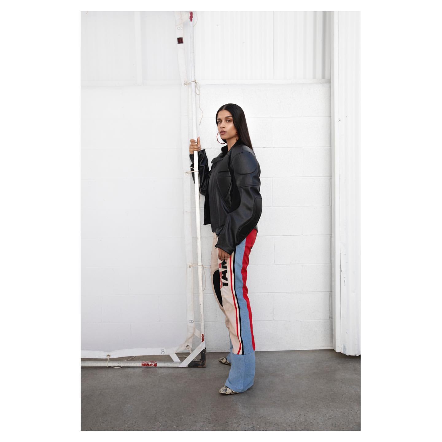 Stylish woman posing indoors in a motorcycle jacket and colorful racing pants, leaning on an adjustable support pole, looking off-camera.