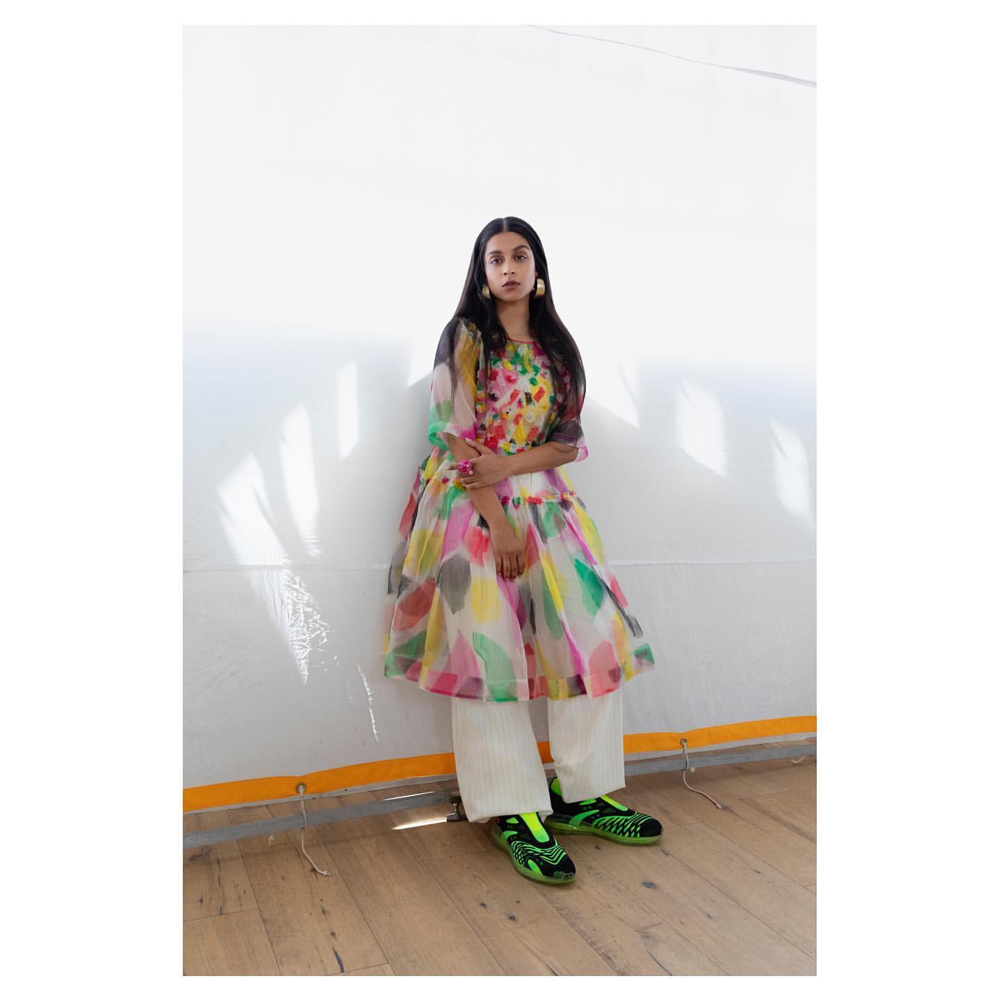 A person in a colorful, floral patterned midi dress stands against a white wall wearing white pants and bright green sneakers in a studio.