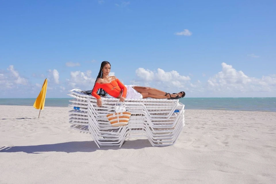 Woman in orange top and white skirt lies on a stack of beach chairs with a straw bag on a sunny beach by the ocean.