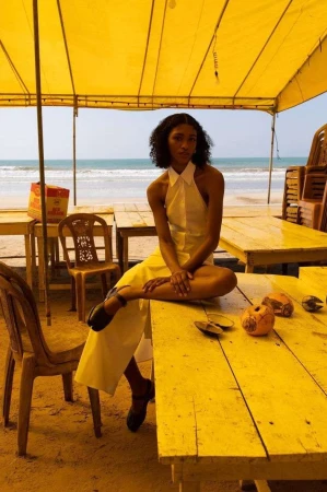 Model seated on a yellow table under a yellow canopy by the beach wearing a white halter dress