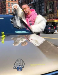 J Balvin wearing a white faux fur coat and pink hoodie dusting a luxury car windshield on a New York street by Nick Sethi.