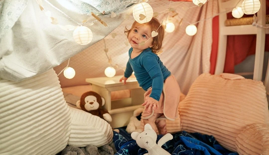 A toddler in a makeshift indoor tent, illuminated by string lights, creates a cozy, playful atmosphere filled with soft pillows and stuffed animals.