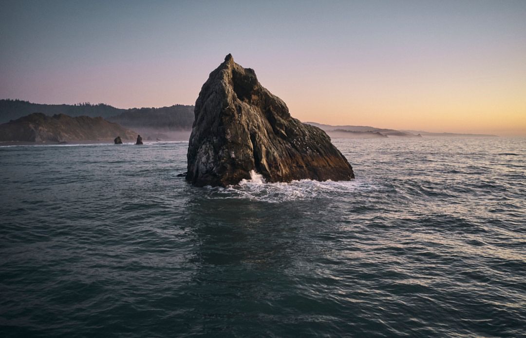 Large rock formation rises from the ocean under soft light, waves crashing at its base, with distant shore and pastel sky at dusk.