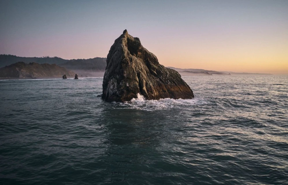 Large rock formation rises from the ocean under soft light, waves crashing at its base, with distant shore and pastel sky at dusk.