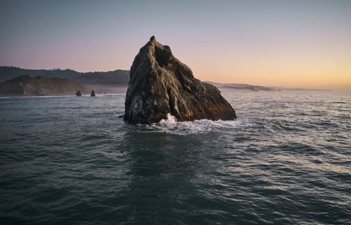 Large rock formation rises from the ocean under soft light, waves crashing at its base, with distant shore and pastel sky at dusk.
