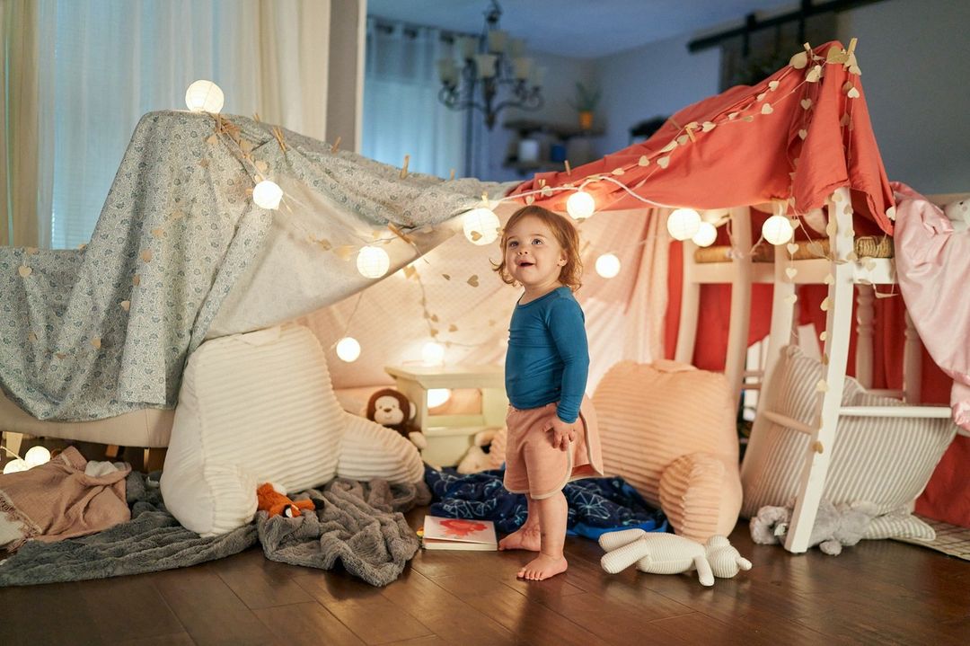 A young child stands in front of a blanket fort decorated with string lights in a cozy indoor living room setting at home.