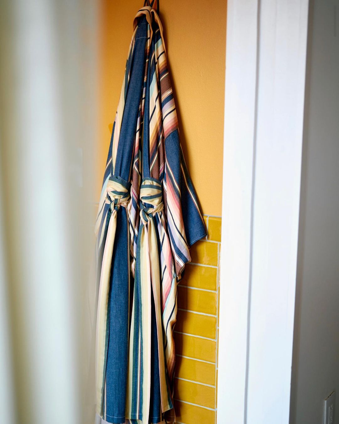 Striped robe hanging on a hook against a yellow tiled wall, next to a white door frame and white shower curtain.