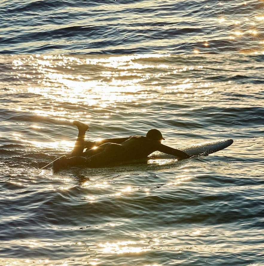 A surfer paddles on their stomach in dark water, sunlight reflects and glistens off the ocean's surface at golden hour.