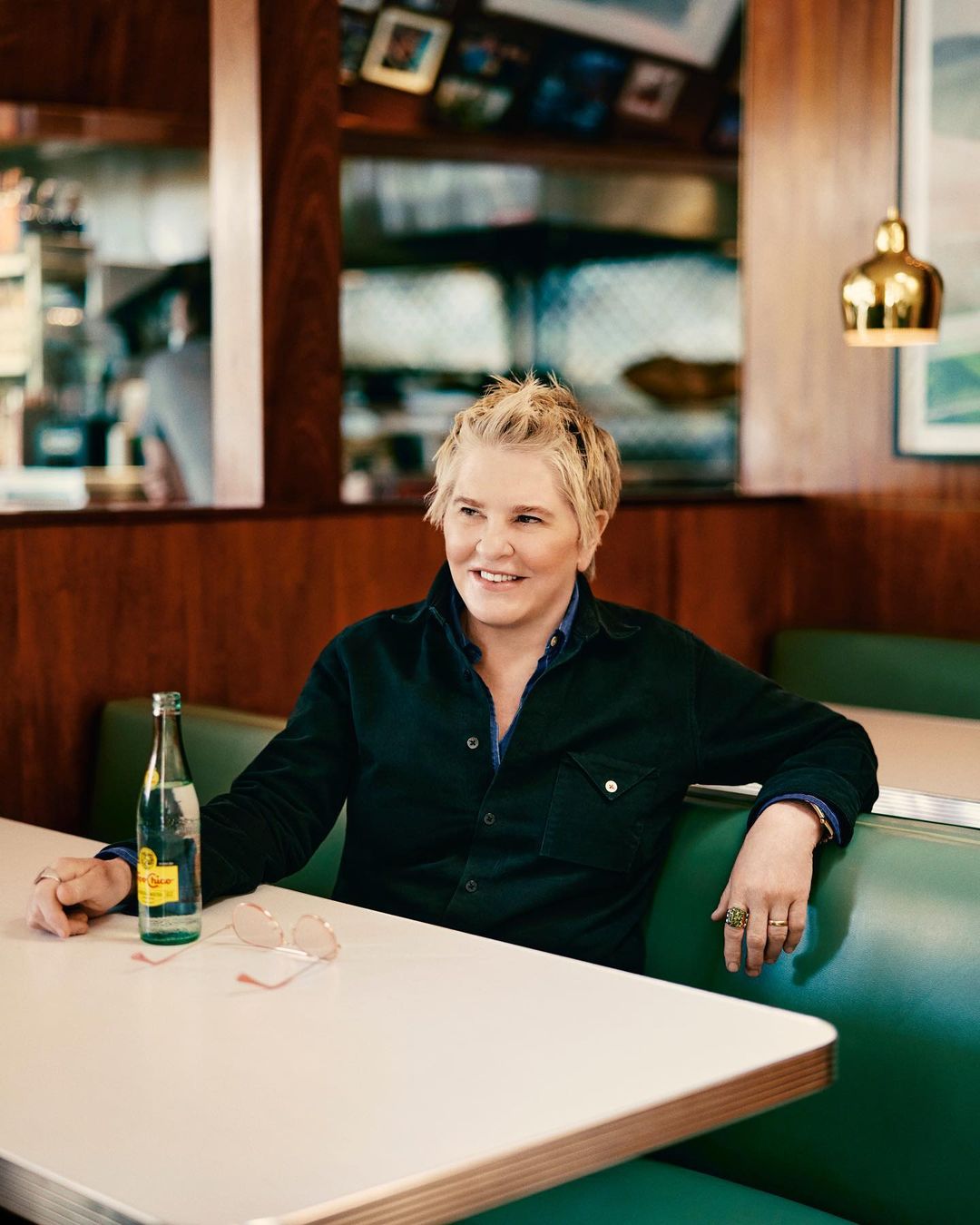 Adult woman seated at a booth in a restaurant with water bottle and sunglasses on table. Interior with wood paneling visible.
