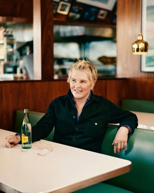 Adult woman seated at a booth in a restaurant with water bottle and sunglasses on table. Interior with wood paneling visible.