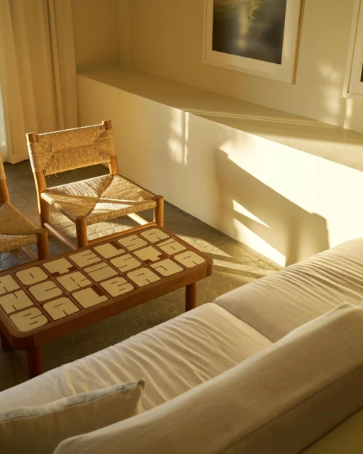 Living room interior with straw chairs, a low table with letter tiles, and a cream-colored sofa, illuminated by sunlight.