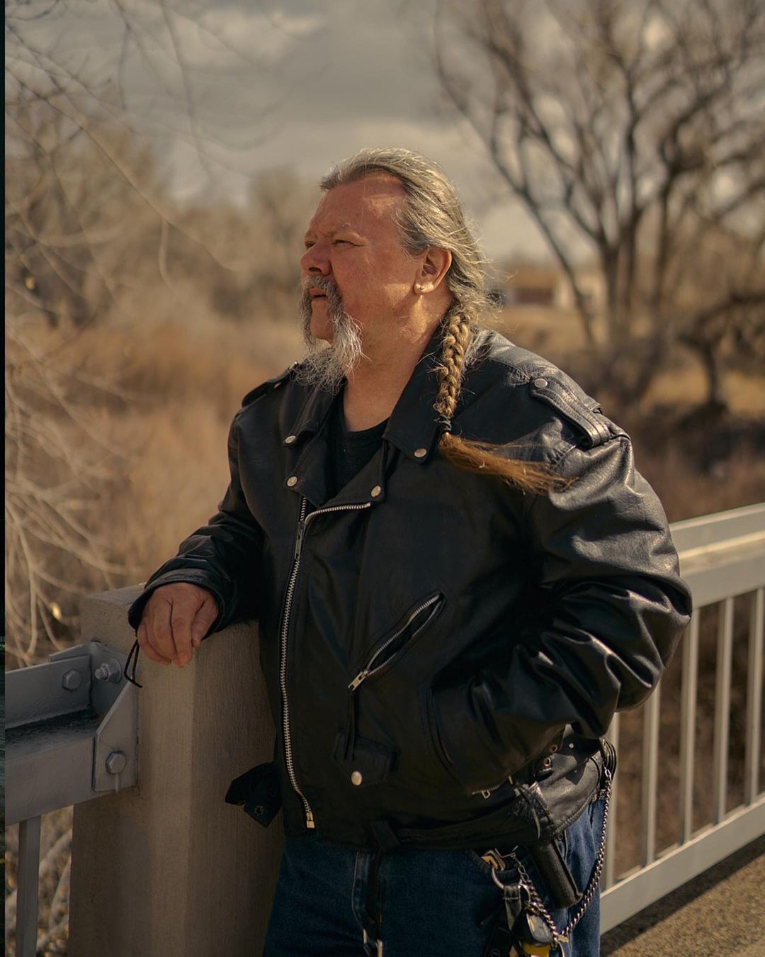 An older adult man with long braided hair and a leather jacket stands on a bridge overlooking a dried riverbed in the daylight.