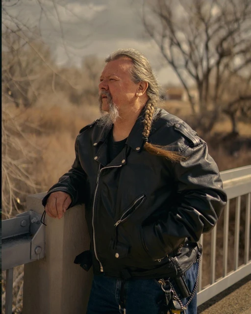 An older adult man with long braided hair and a leather jacket stands on a bridge overlooking a dried riverbed in the daylight.