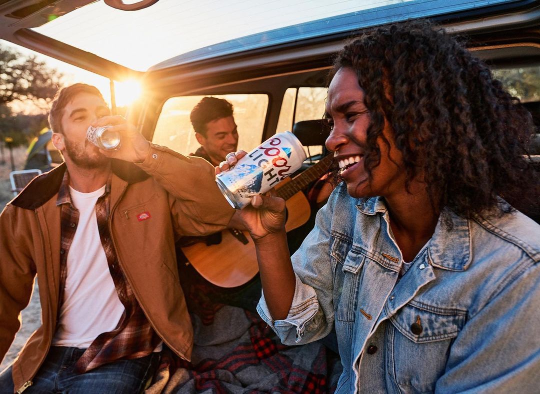 Three young adults enjoy Coors Light beer and play acoustic guitar outdoors with bright sunset light streaming in from the open car hatch.