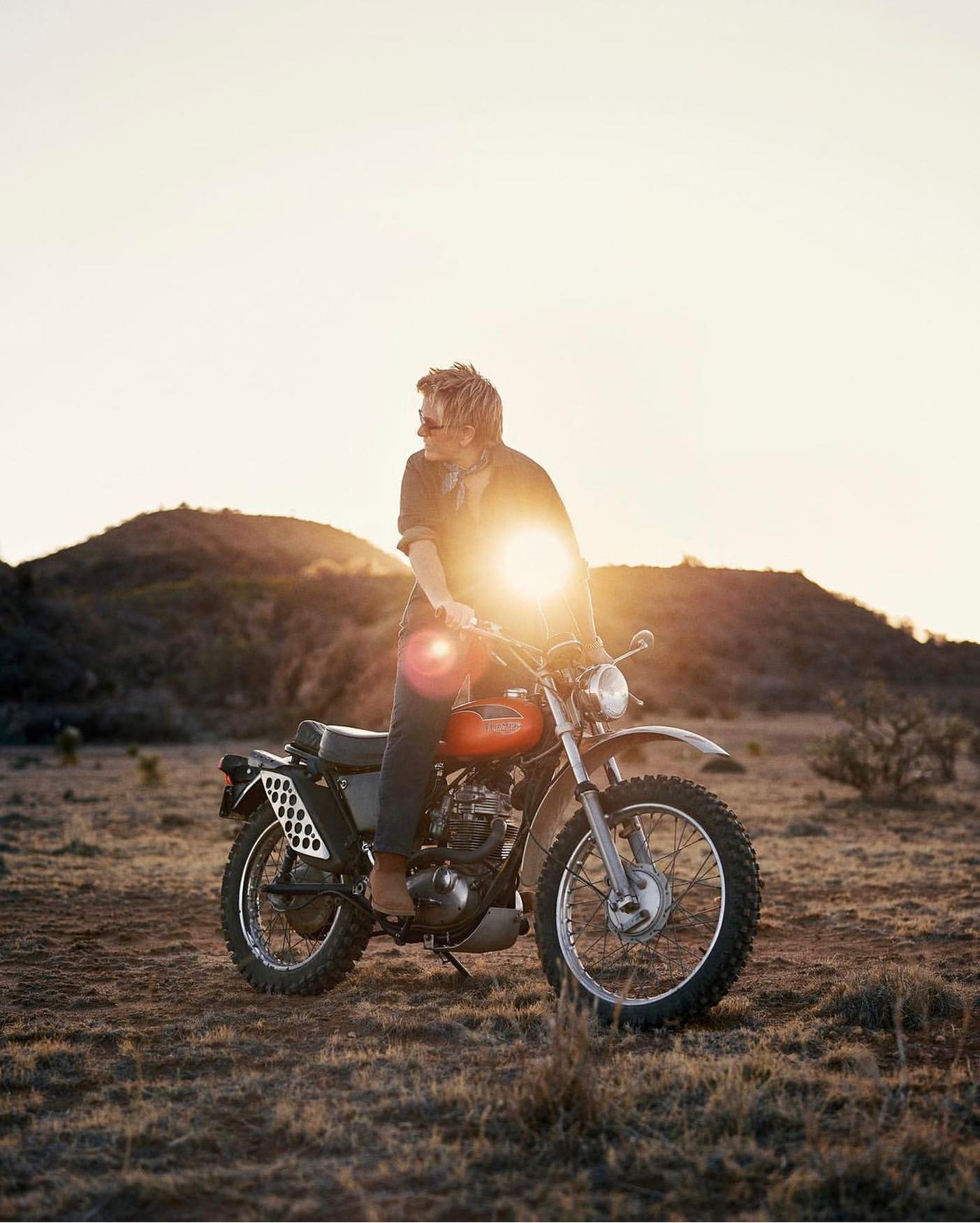 Adult male rides a vintage orange and silver motorcycle on a grassy plain at sunset, with rolling hills in the background.