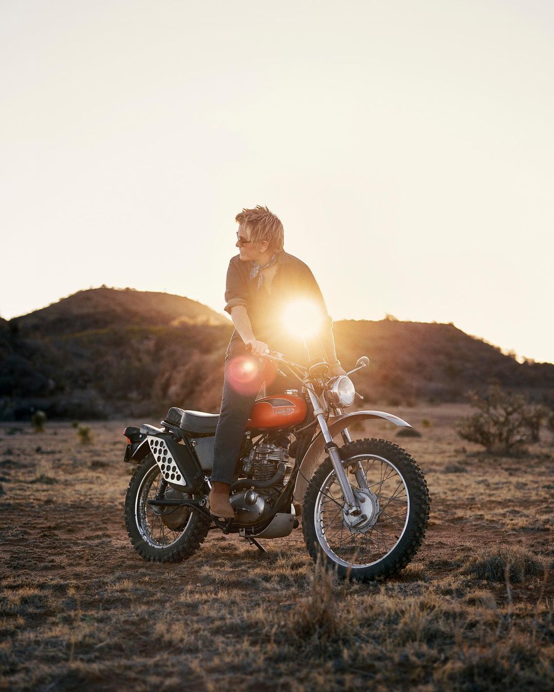 A man riding a vintage motorcycle through a rural, grassy landscape under a bright, golden sunset with hills in the background.