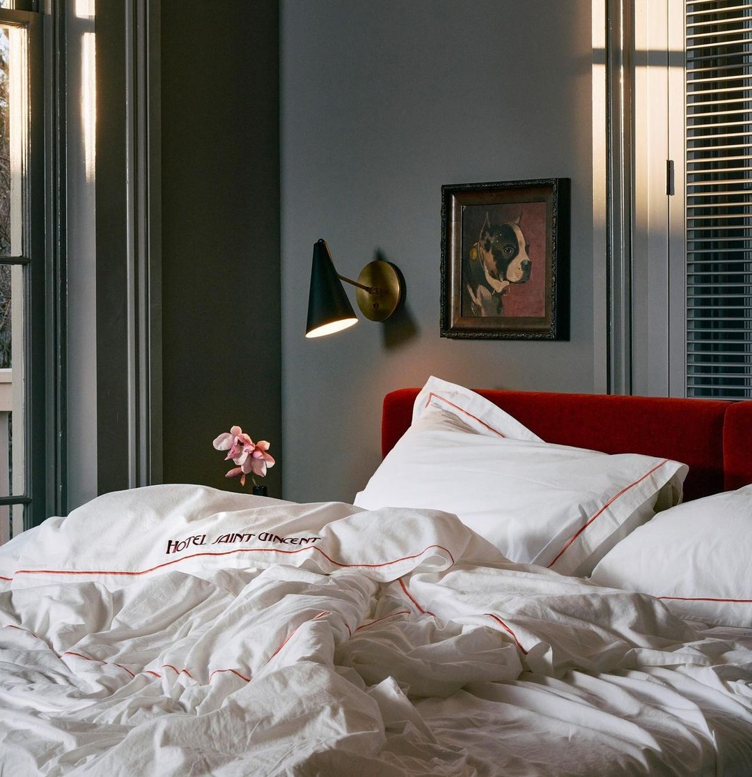 Interior view of a hotel room bed with white linens, a red headboard, a wall-mounted light fixture, and a framed dog portrait.