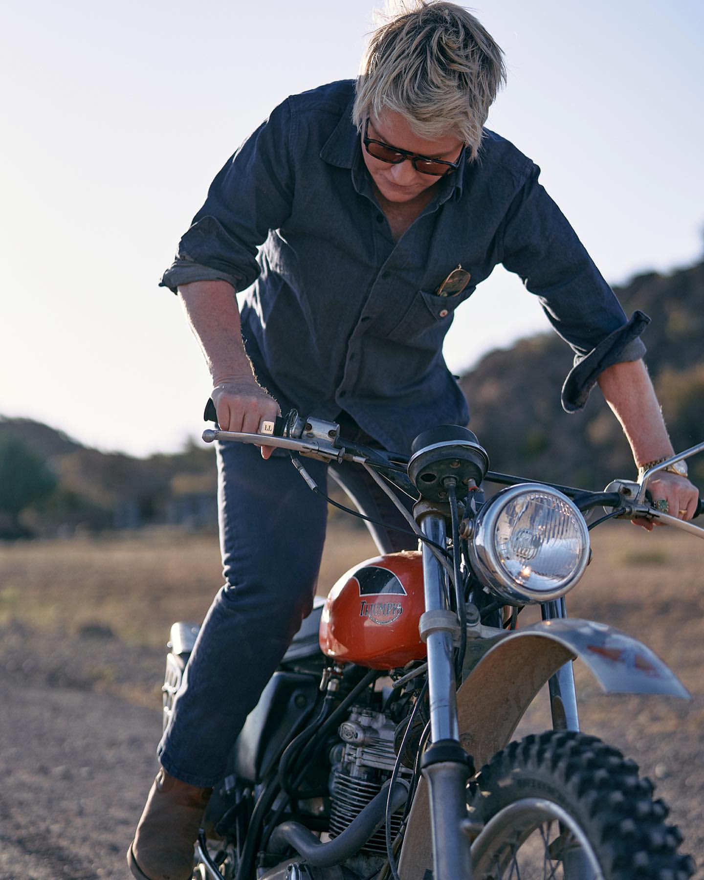 Adult wearing a denim shirt and sunglasses sits on a vintage orange and silver motorcycle in a desert landscape on a sunny day.