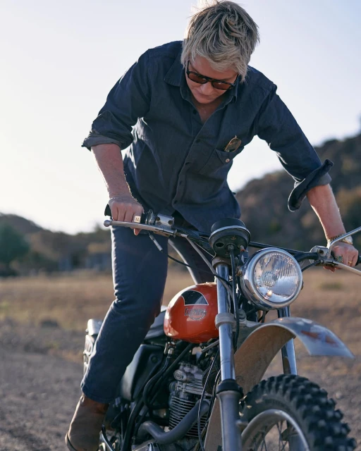 Adult wearing a denim shirt and sunglasses sits on a vintage orange and silver motorcycle in a desert landscape on a sunny day.