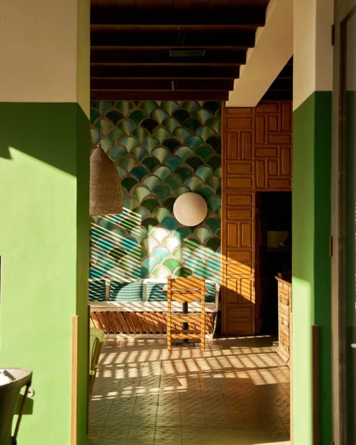 Interior view showcasing a tiled wall with a circular lamp, a wooden bench, and sunlight streaming through blinds, creating strong shadows.