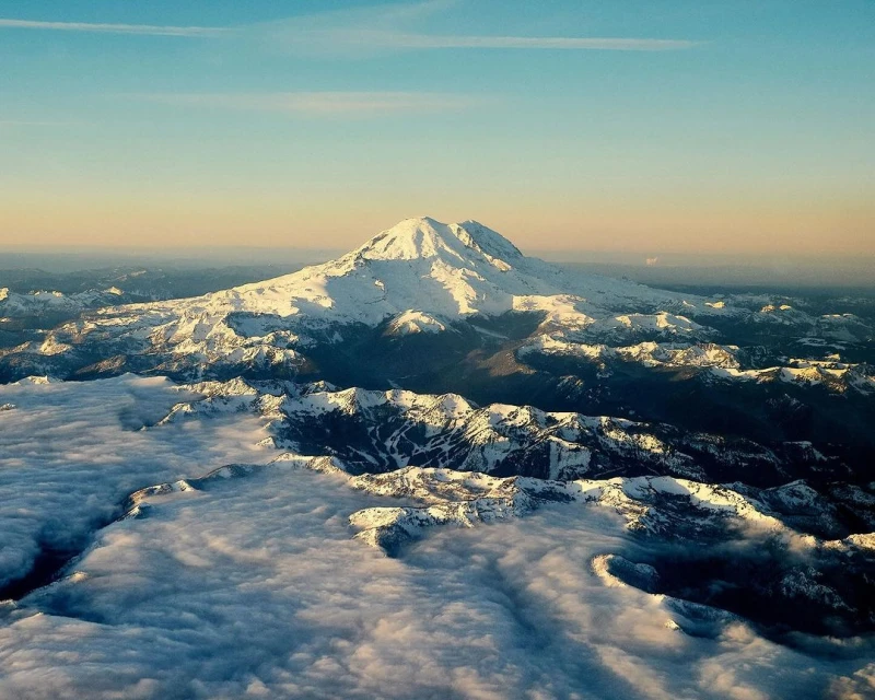 Aerial view of snow-capped mountain peak, mountain range, and low-lying clouds under a gradient blue and orange sky at daytime.