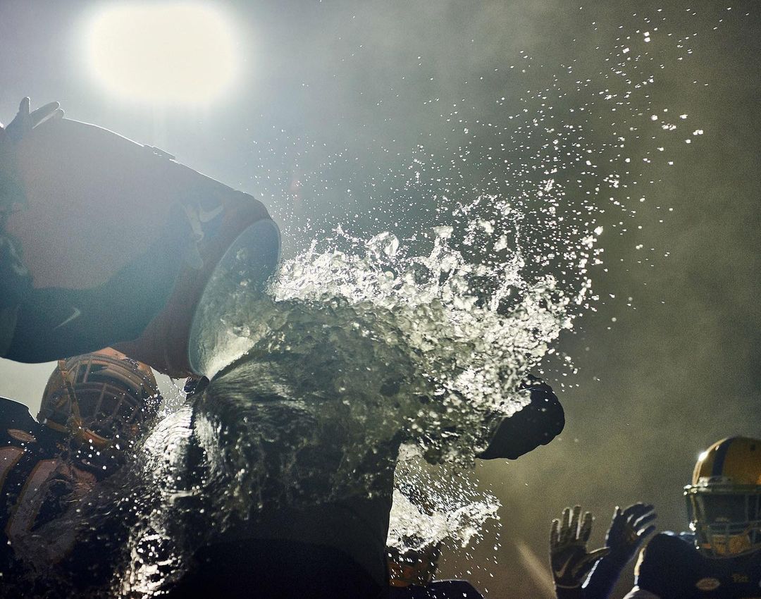 American football players celebrate a victory by dousing a teammate with water; helmets and uniforms visible in a dark, bright environment.