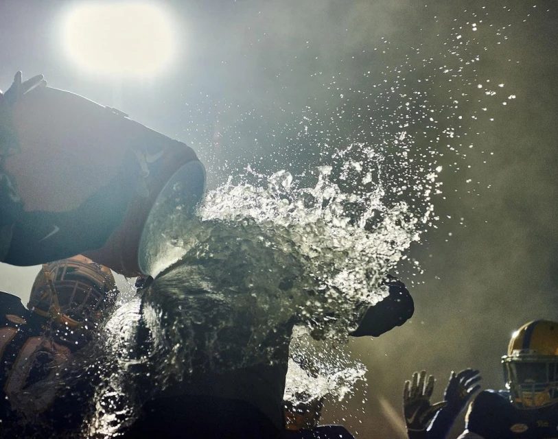 American football players celebrate a victory by dousing a teammate with water; helmets and uniforms visible in a dark, bright environment.