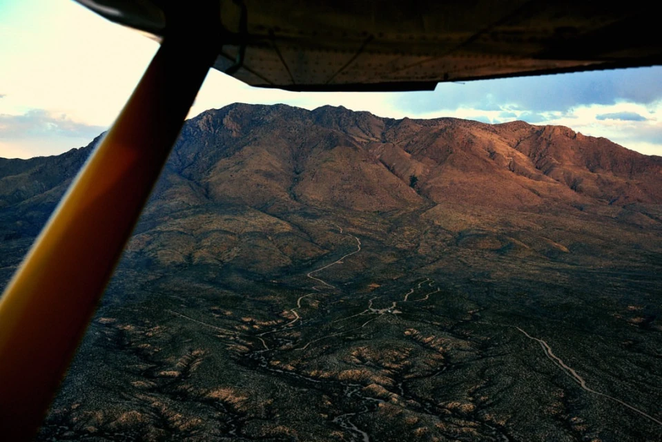 Aerial view of a desert mountain range with winding dirt roads, seen through the window of a small airplane on a sunny day.