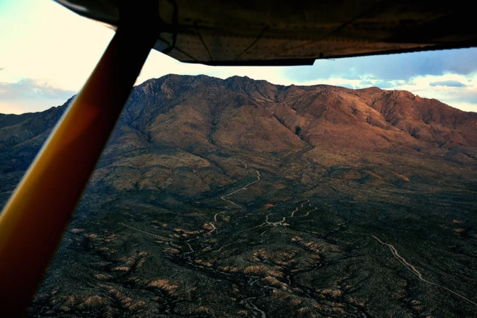 Aerial view of a desert mountain range with winding dirt roads, seen through the window of a small airplane on a sunny day.