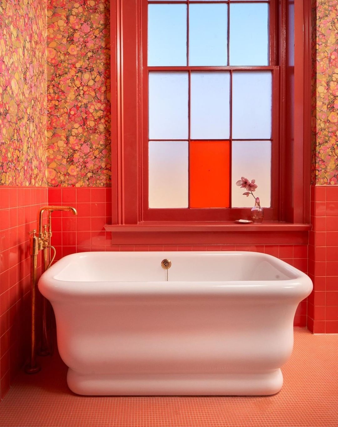 Bathroom interior featuring a white bathtub, red tile walls, floral wallpaper, and a window with an orange-tinted pane.