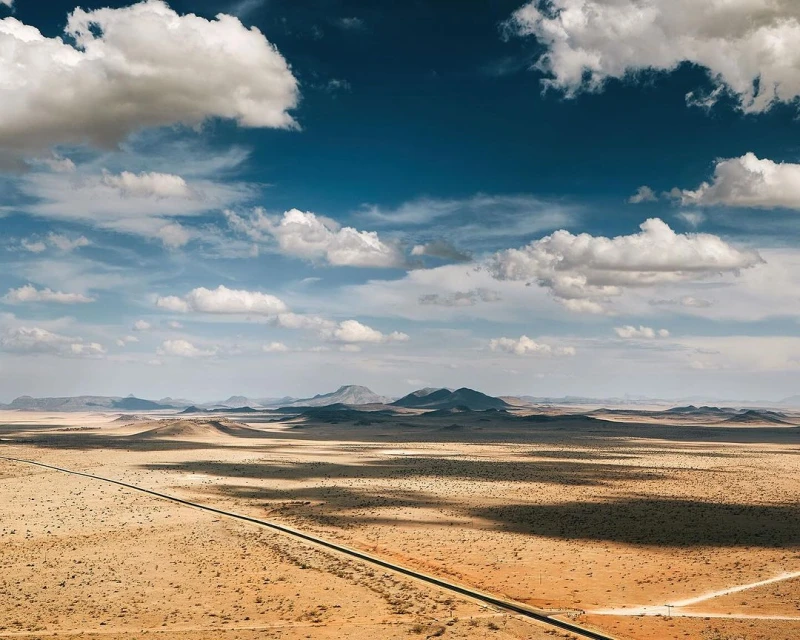 Aerial view of an arid landscape with distant mountains under a partly cloudy blue sky, featuring a road stretching into the distance.