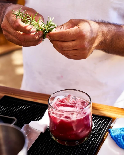 Bartender prepares a vibrant red cocktail, garnishing it with fresh rosemary sprigs over a bar top with ice-filled glass and garnish.