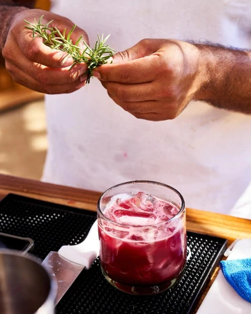 Bartender prepares a vibrant red cocktail, garnishing it with fresh rosemary sprigs over a bar top with ice-filled glass and garnish.